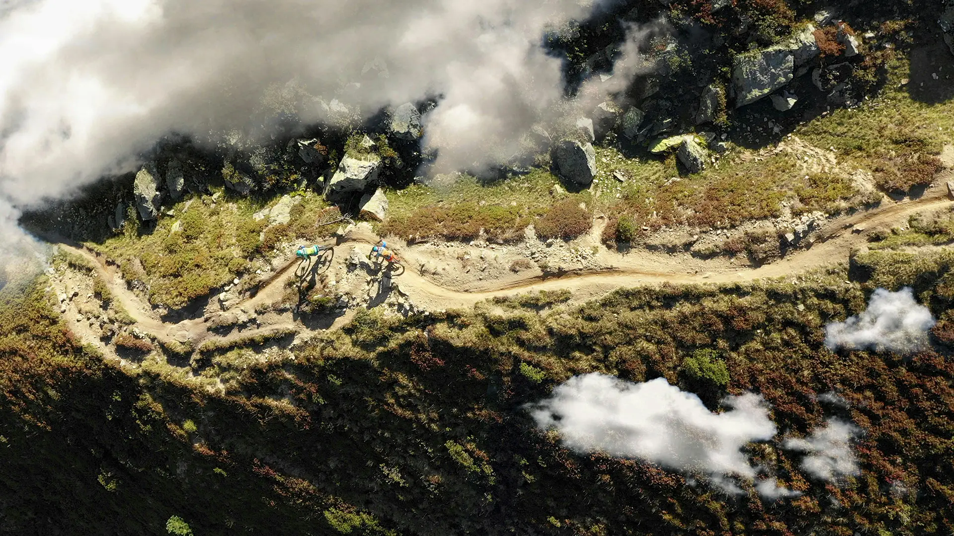 Aerial view of mountain bikers riding along a dirt trail surrounded by rocky terrain and clouds.