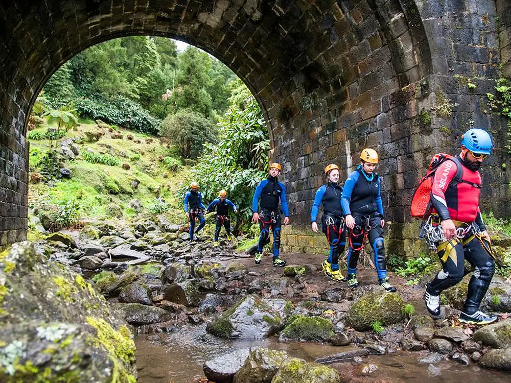 Group of individuals in canyoning gear walking under a stone bridge in a natural setting.