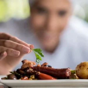 A chef adding a garnish to a plated dish featuring meat, vegetables, and a potato.