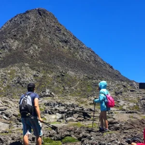 Two hikers and a person taking a photo while approaching Pico Mountain under a clear blue sky.