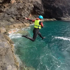 Individual in a wetsuit and helmet jumping off a rocky ledge into the ocean water in Madeira.