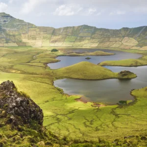 Individual standing on a rock with arms raised, overlooking a green landscape and a lake.