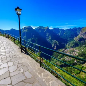 Curved stone pathway with a railing overlooking a mountainous landscape and a village below.