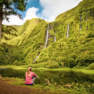 Individual sitting on the shore of a lake, with waterfalls cascading down a green mountain in the background.