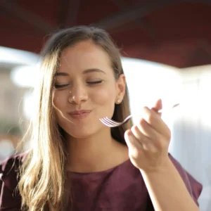 Woman with long hair smiling while eating with a fork outdoors under an umbrella.