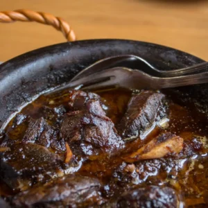 A clay pot filled with cooked meat pieces and sauce, accompanied by a fork resting on the side.