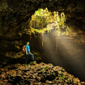 Individual standing on rocks inside a cave, illuminated by natural light from above.