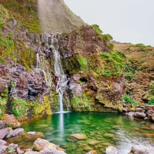 A waterfall flowing down rocky terrain into a clear green pool surrounded by moss and vegetation.