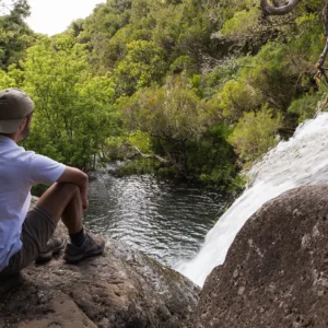Individual sitting on a rock near a waterfall, surrounded by greenery and water.