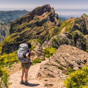 Person with a backpack walking on a rocky trail in a mountainous area with greenery.