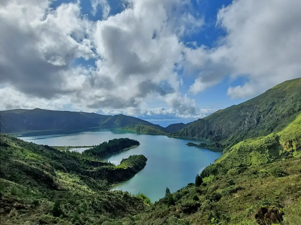 A panoramic view of Lagoa do Fogo, a lake surrounded by green hills and mountains under a cloudy sky. Available in Azores / Açores.