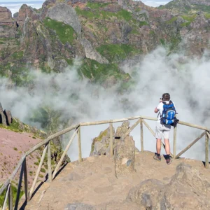 Individual standing on a wooden platform with a mountainous landscape and clouds in the background.