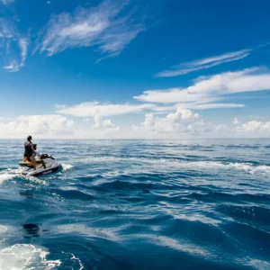 Person riding a jet ski across calm ocean waters under a blue sky with clouds.