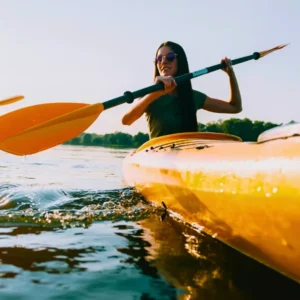 Individual paddling a yellow kayak on calm water with a paddle in hand and sunglasses on.