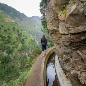 Individual walking on a narrow path beside a water channel in a mountainous landscape.
