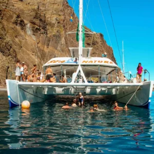A catamaran boat with passengers on board in the water near rocky cliffs in Madeira.