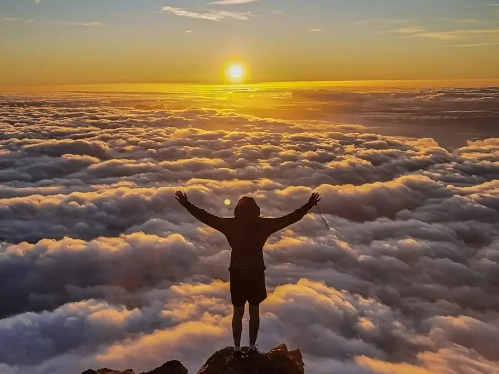 Individual with arms outstretched standing on a mountain peak above the clouds during sunset.