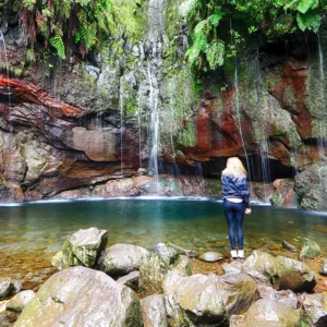A woman standing by a pool of water in front of a waterfall surrounded by rocks and greenery.