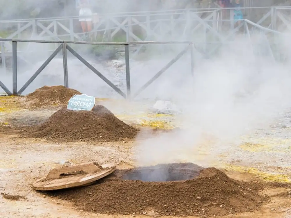 Geothermal feature with steam rising from a hole in the ground at Furnas Lake, surrounded by mounds of earth.