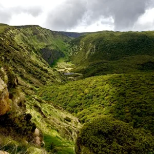 A panoramic view of a lush green valley surrounded by hills under a cloudy sky.
