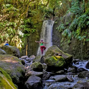Individual balancing on rocks in a stream near a waterfall surrounded by lush greenery.